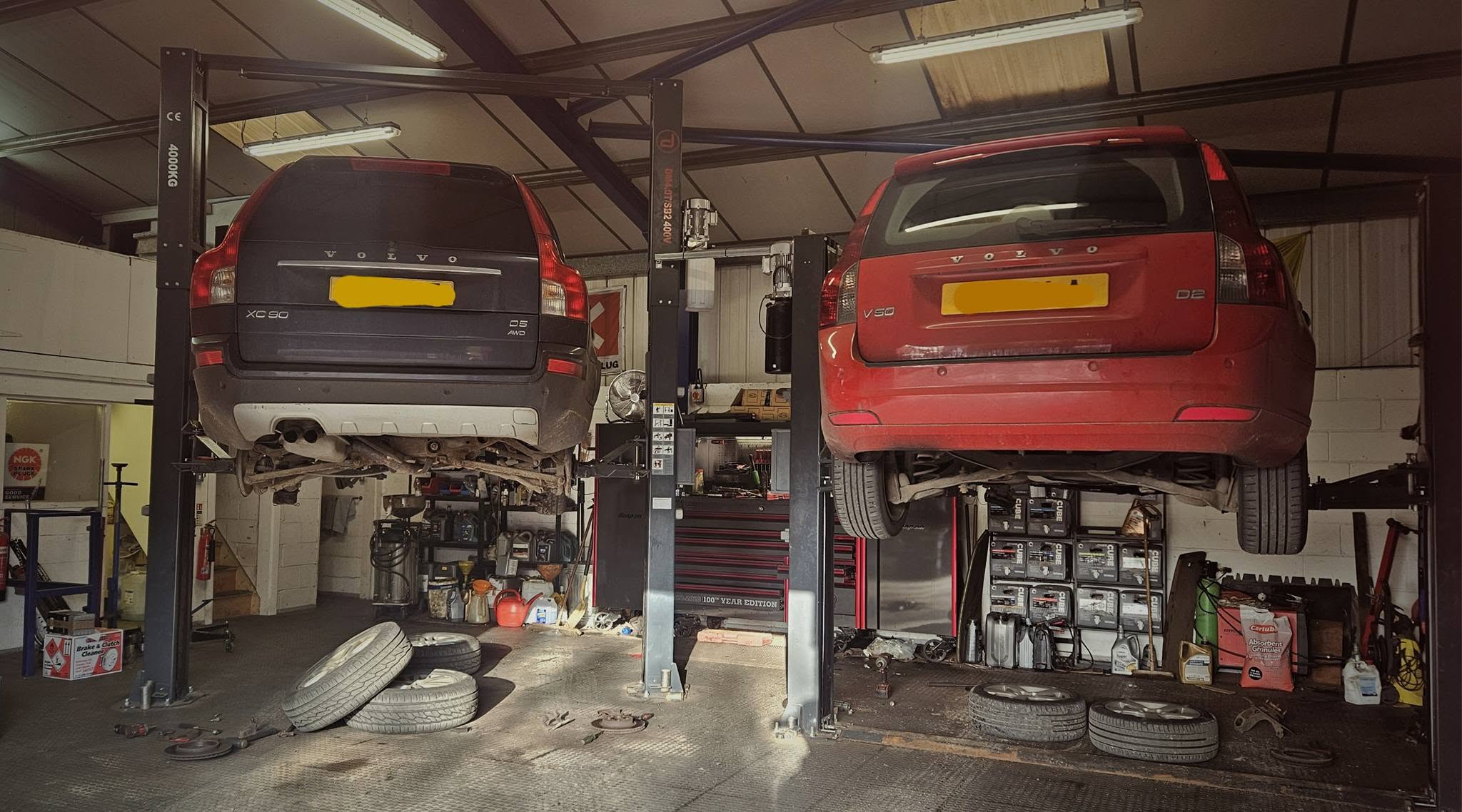A grey Volvo XC90 and a red Volvo raised side by side on two lifts with wheels and tyres on the floor