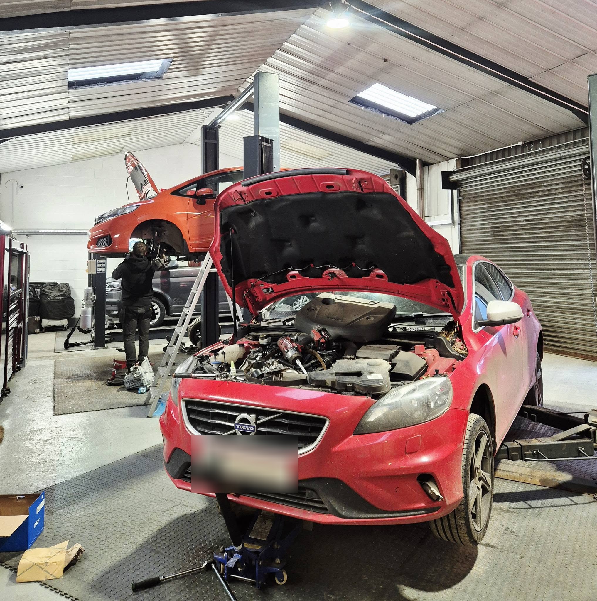 A red Volvo V40 with its bonnet up and engine parts off, a Ford Fiesta raised on the lift behind and a mechanic working on it