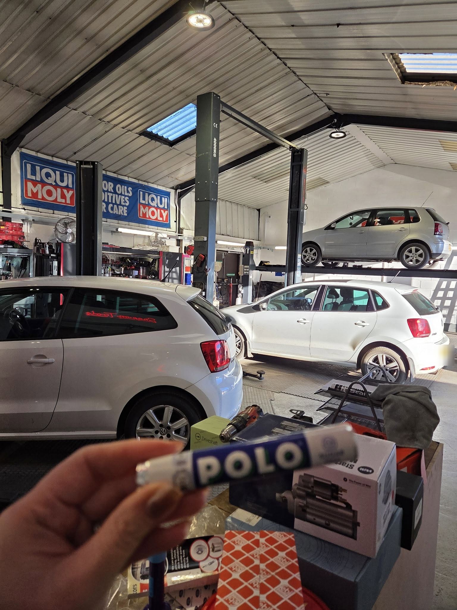 A tube of Polo mints held up in front of two white VW Polos in the workshop, one on a lift