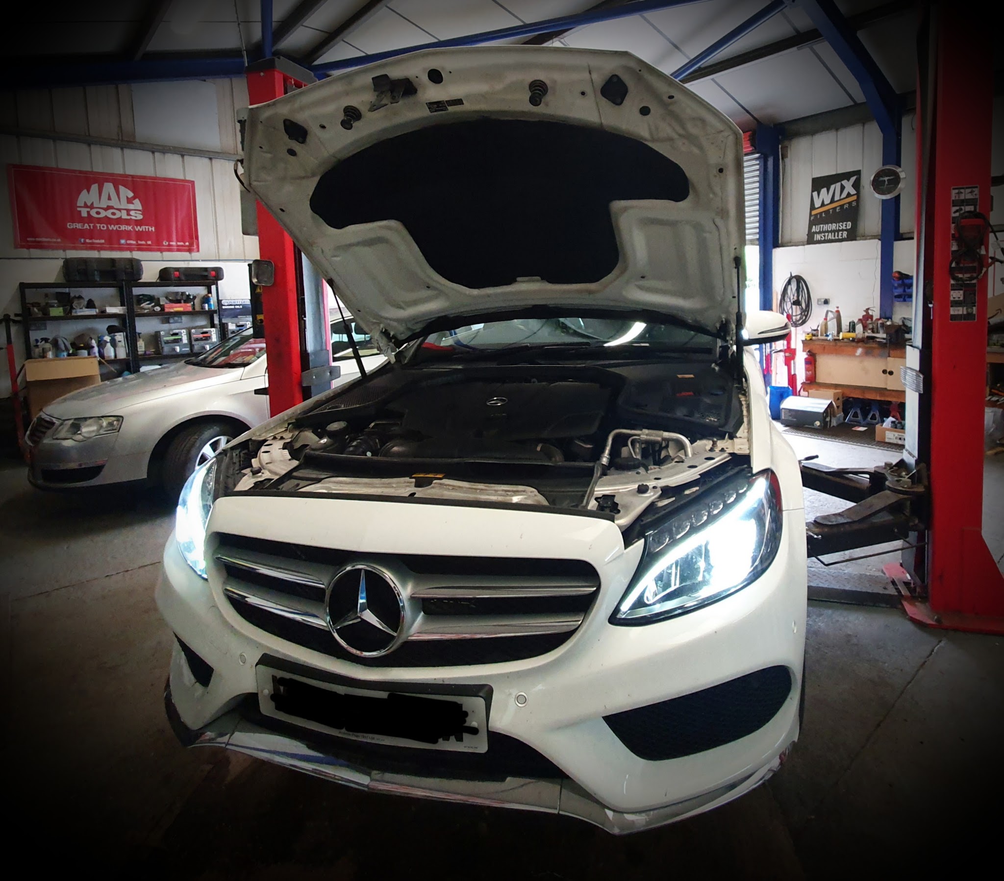 A white Mercedes C-Class in the workshop with its bonnet up and headlights on