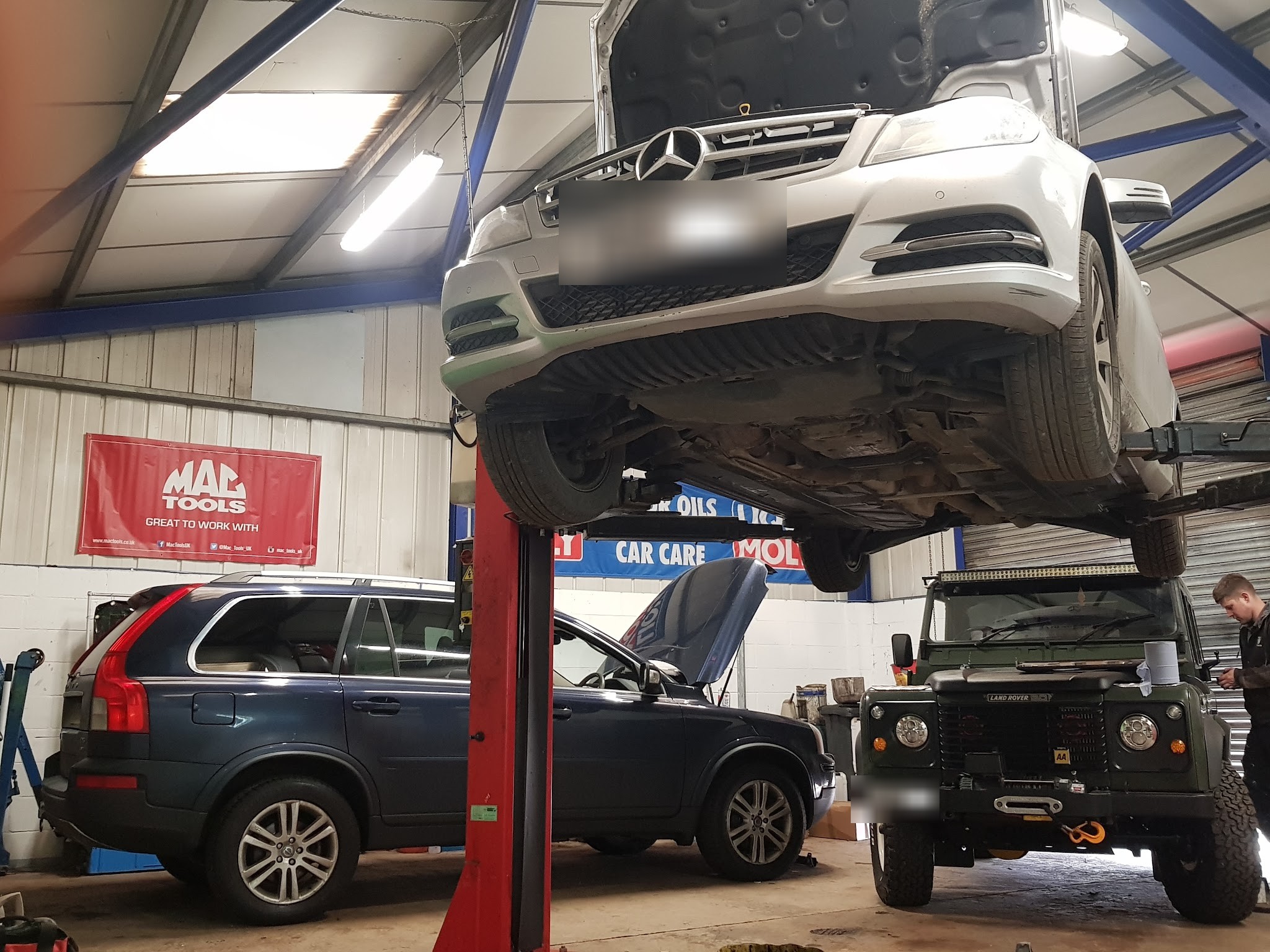 A silver Mercedes up on a lift with a Volvo XC90 and a Land Rover Defender parked underneath in the workshop